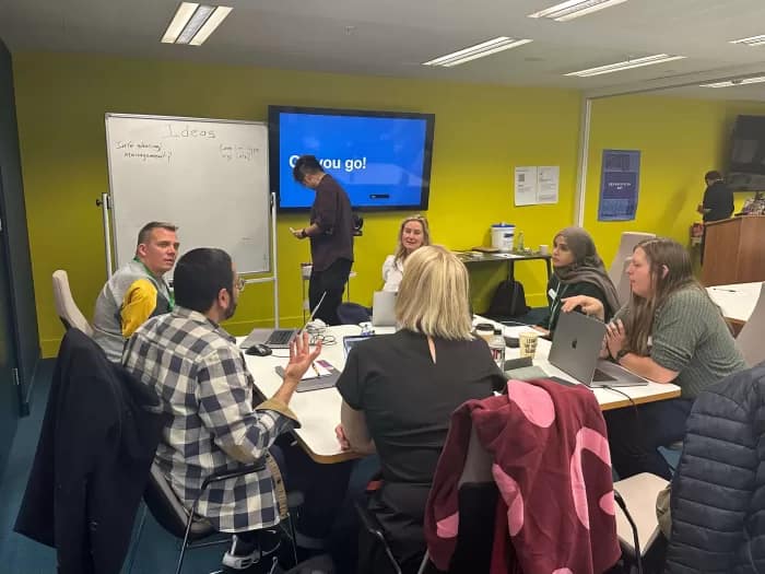 Design System Hackday attendees gather around a table and board to think of ideas.